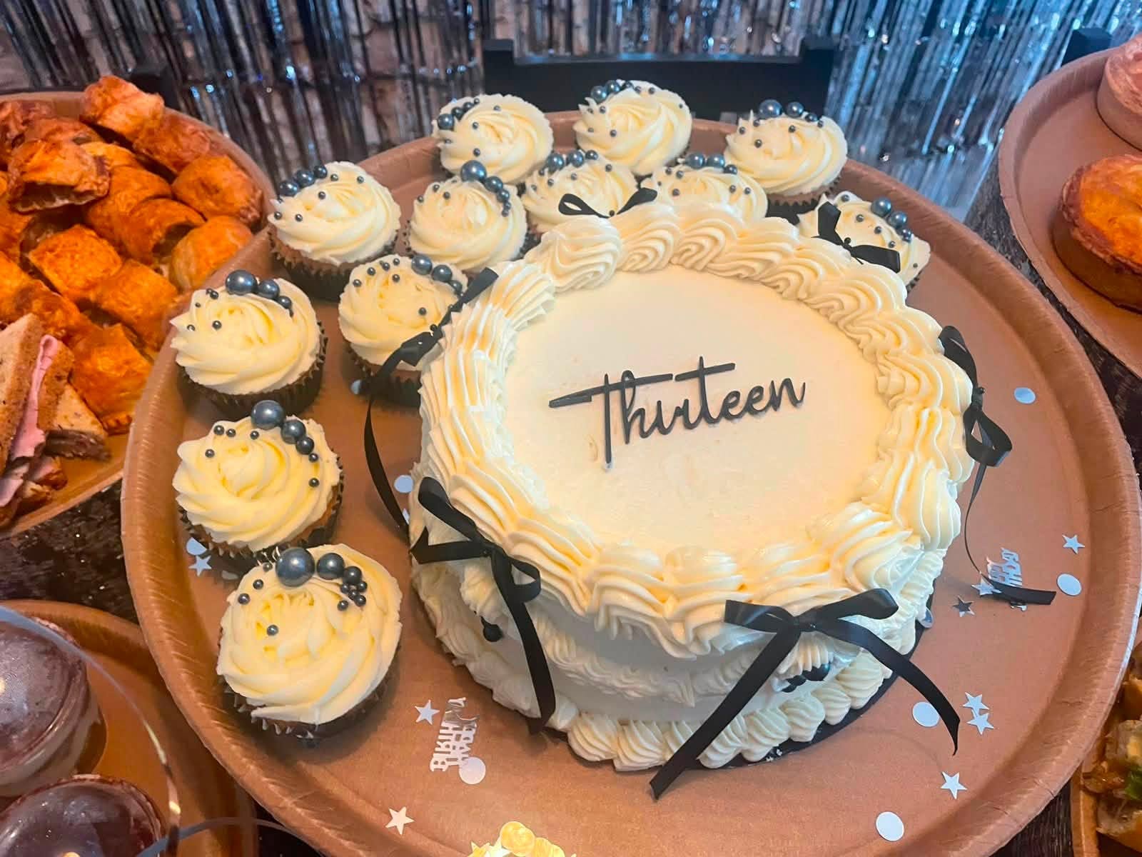 A white buttercream thirteenth birthday cake piped with ruffles and tied with black bows, surrounded by cupcakes topped with silver pearls