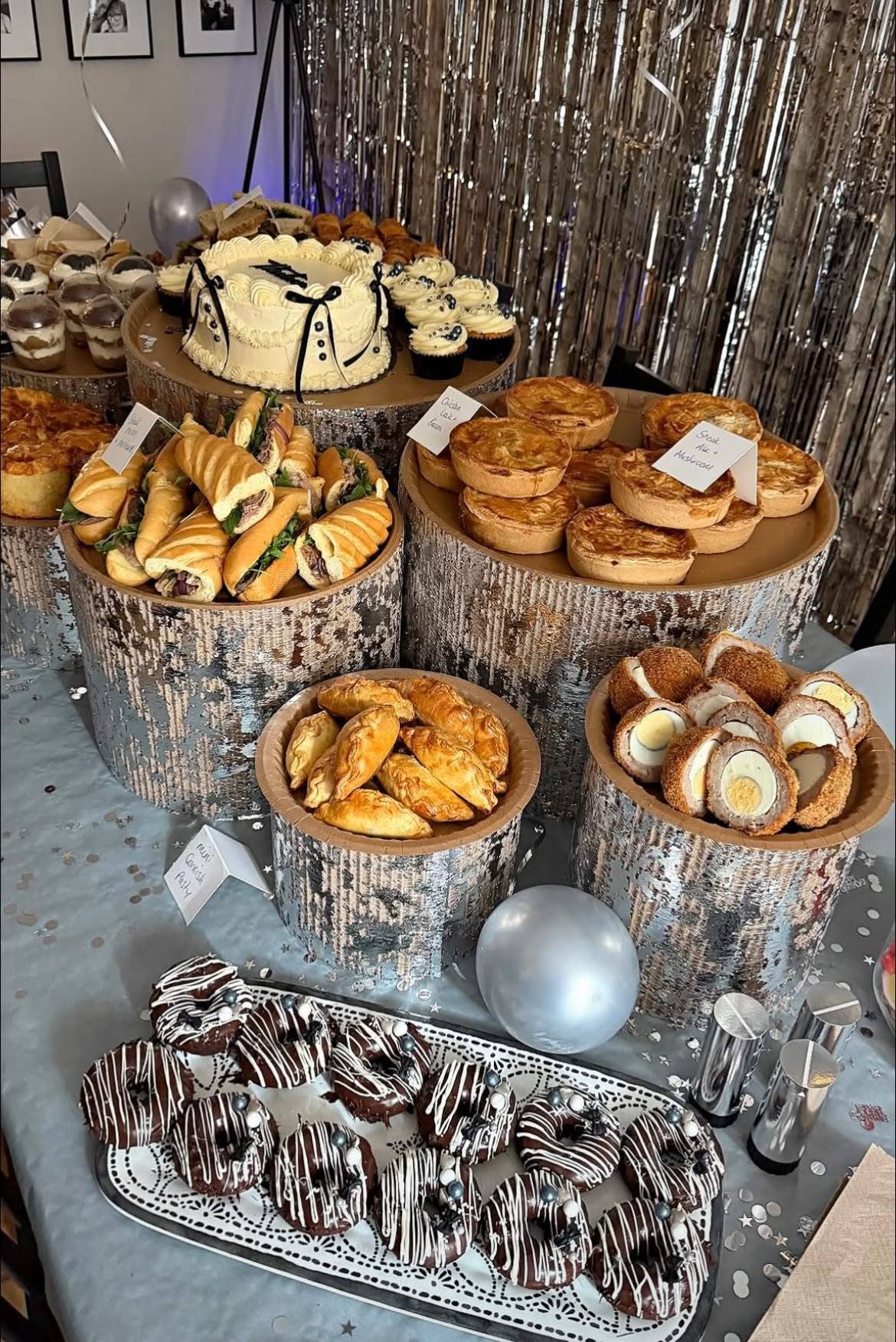 A party table with a white celebration cake, baguettes, quiches, pasties, scotch eggs and chocolate iced doughnuts