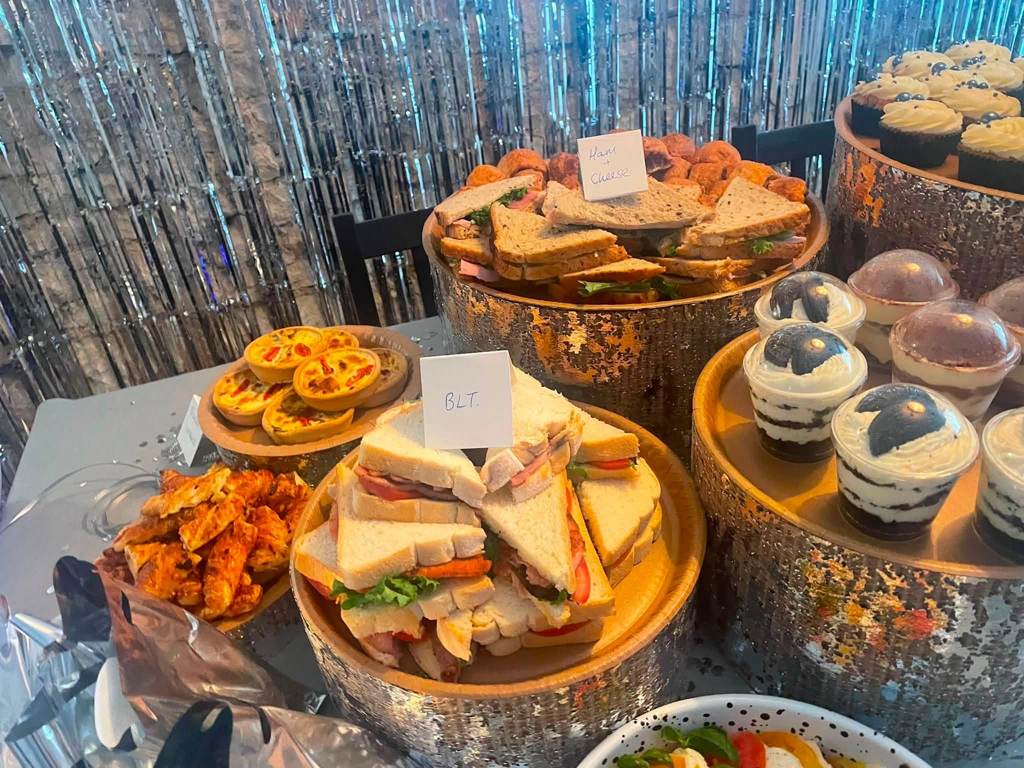Wooden stands holding ham and cheese sandwiches, BLT sandwiches, quiches, spiced chicken and cookies and cream dessert pots