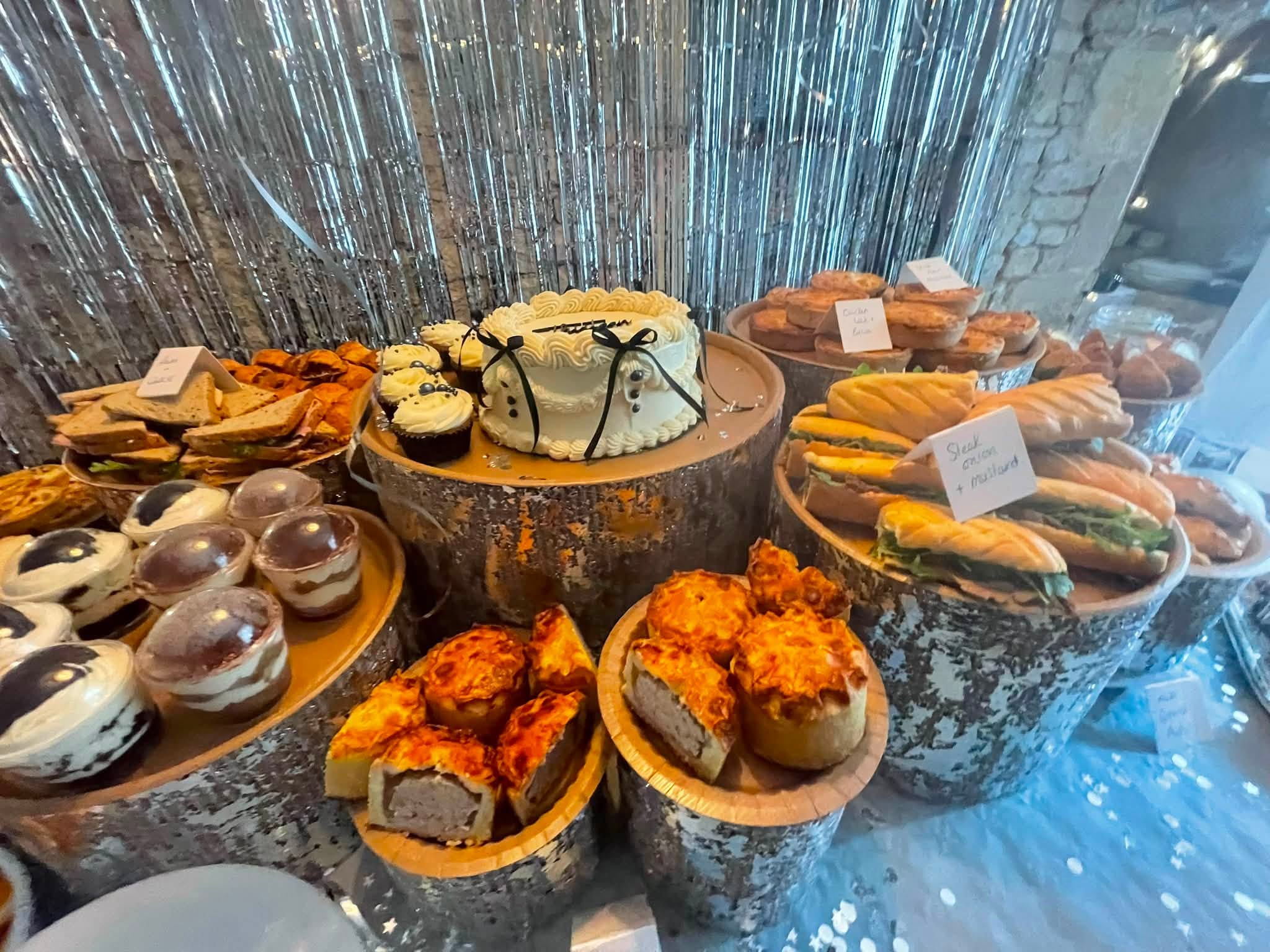 A grazing spread centred on a white buttercream celebration cake surrounded by pork pies, baguettes, quiches and dessert pots