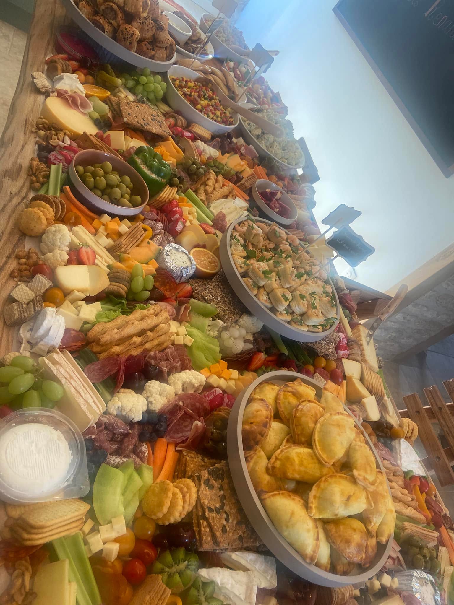 A long wooden table covered end to end with cheeses, charcuterie, crackers, olives, fresh fruit, empanadas and vol-au-vents