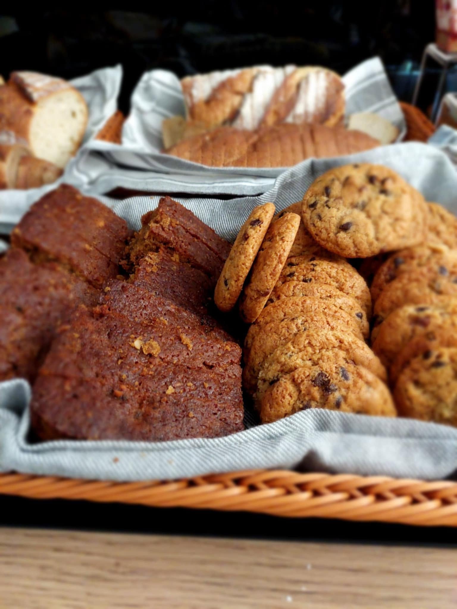A linen lined basket of home baking with slices of banana loaf, chocolate chip cookies and biscuits, with fresh bread behind