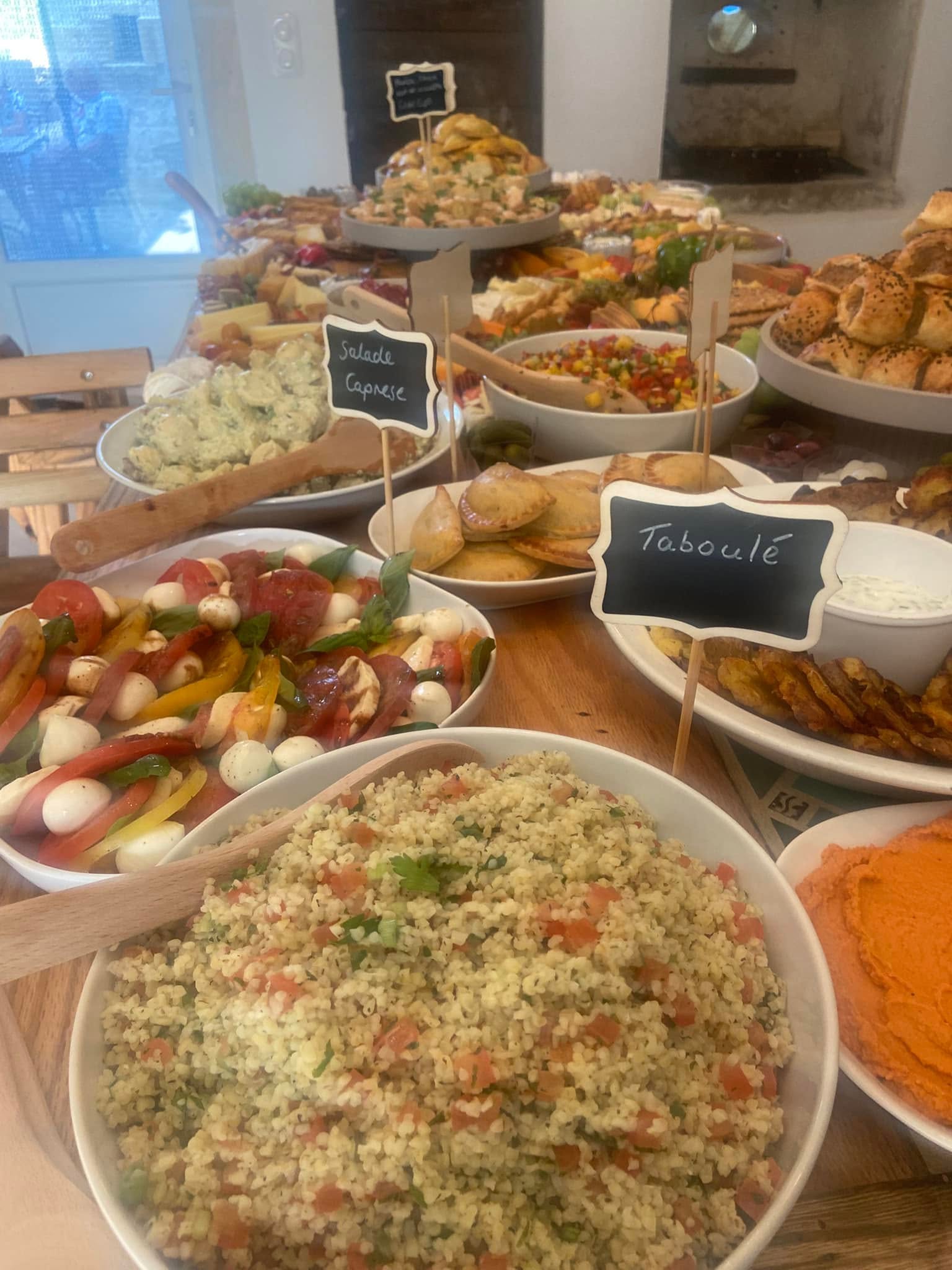 A long buffet table in a home kitchen with chalkboard labelled dishes of caprese salad, tabbouleh, empanadas, sausage rolls and houmous