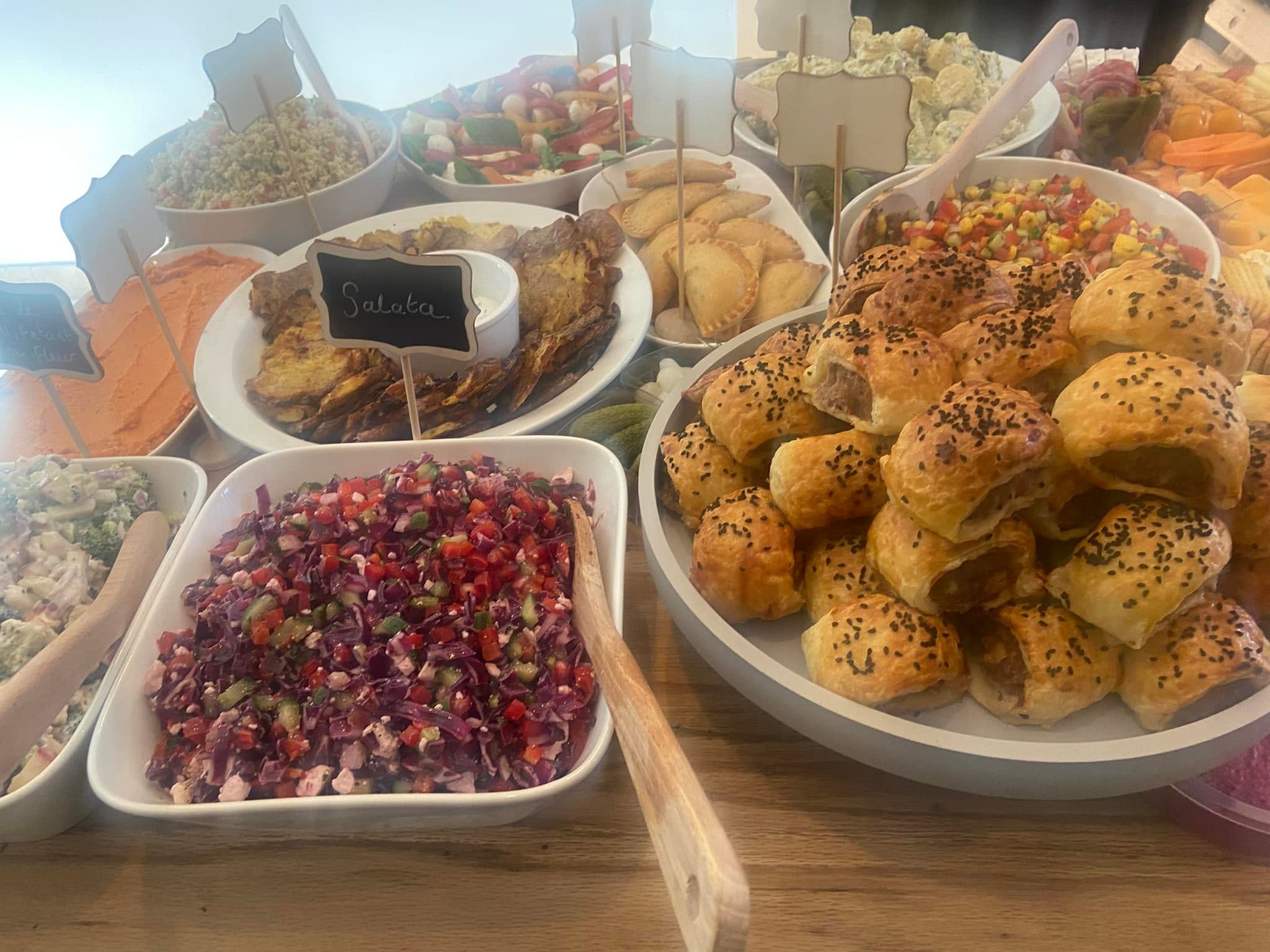 A spread of labelled salads and dips with sausage rolls, empanadas, coleslaw, houmous and couscous