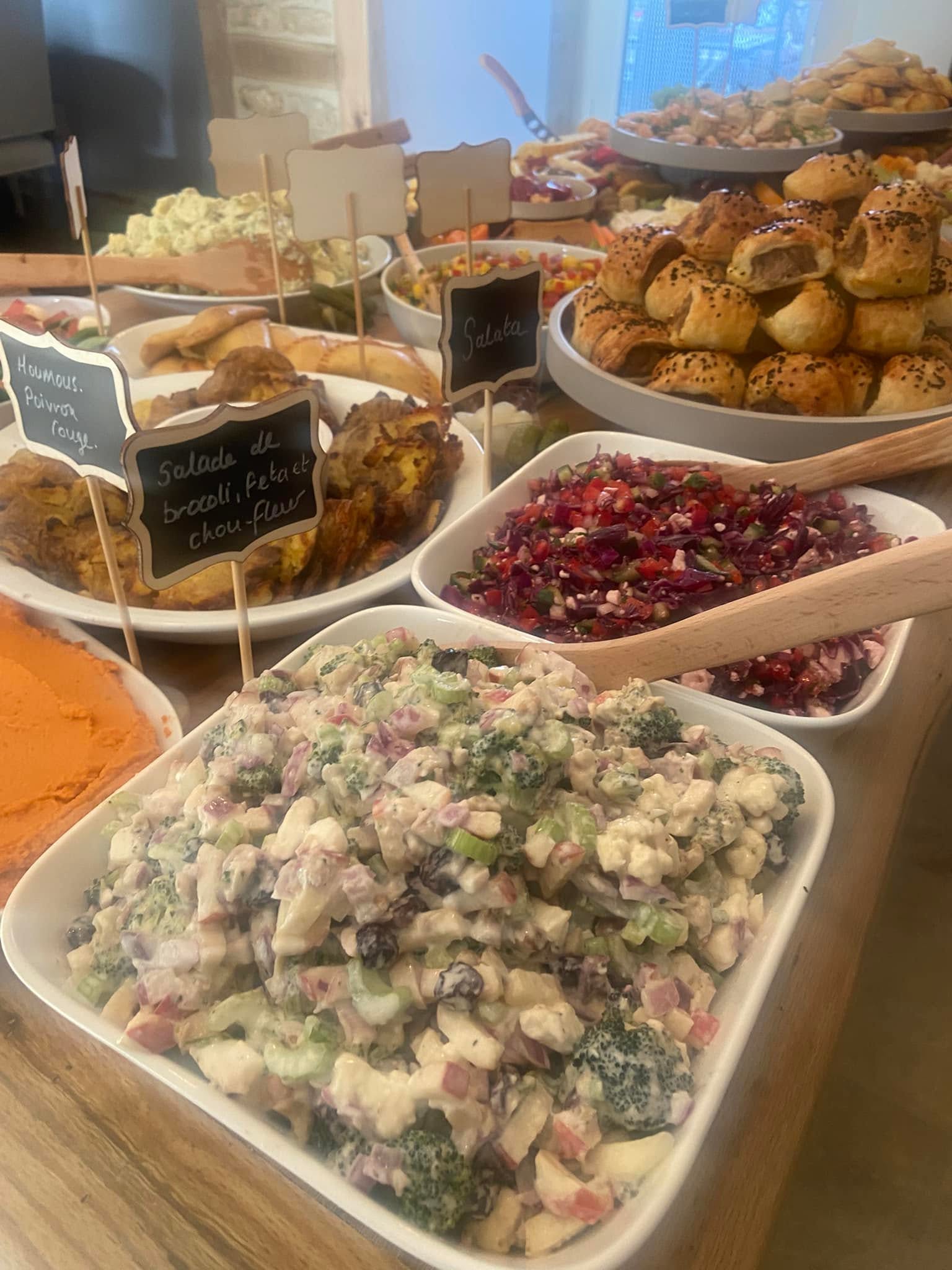 Chalkboard labelled bowls of broccoli and feta salad, red cabbage slaw, houmous and freshly baked sausage rolls